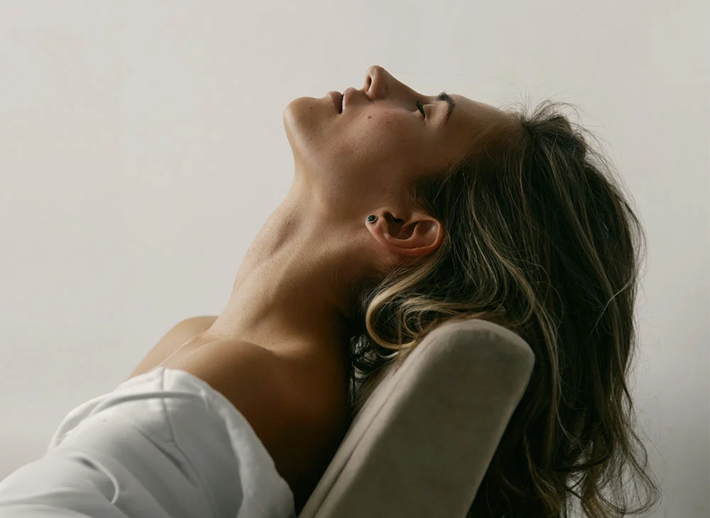 Portrait of a woman with long, wavy light brown hair, reclining on a chair with her eyes gently closed. Her relaxed posture and peaceful expression suggest a moment of quiet contemplation. The soft, diffused lighting and the muted background create a serene and intimate portrait, highlighting her natural beauty and sense of tranquility. - Chin Augmentation in San Clamente, Aliso Viejo and Rancho Santa Margarita, CA