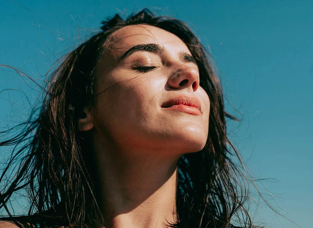 Close-up of a woman with long, windblown dark brown hair against a clear blue sky. Her eyes are closed, and her expression is serene. She has light tan skin with subtle sunlit highlights on her nose and cheeks. The vibrant blue sky provides a contrasting backdrop. - Diamond Glow in San Clamente, Aliso Viejo and Rancho Santa Margarita, CA