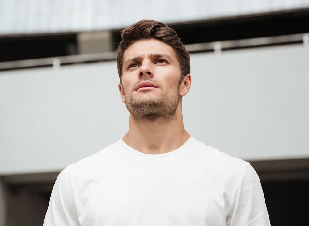 Mid-shot of a man. The man is light-skinned with short, brown hair styled neatly. His gaze is directed upward, and his expression is contemplative or thoughtful. He appears to be in his late twenties or early thirties. He has a short, neatly trimmed beard.  He is wearing a plain, off-white or cream-colored t-shirt.  The background is a light gray or white wall with architectural details, suggesting an interior space, possibly a building or parking garage. The background is out of focus, allowing the man to be the central focus of the image. - Exosomes in San Clamente, Aliso Viejo and Rancho Santa Margarita, CA