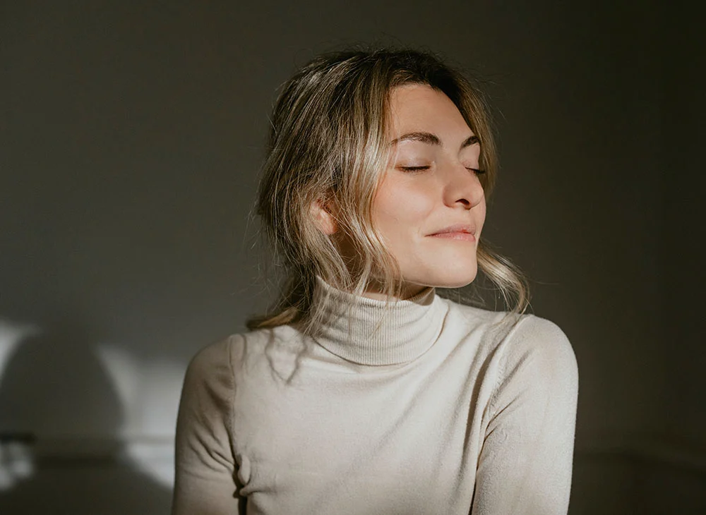 Close-up of a serene woman with light blonde hair and closed eyes, angled slightly to the right. She wears a cream-colored turtleneck sweater, contrasted against a plain dark gray background. Soft natural lighting creates subtle shadows, enhancing the image's mood. - Eyelid Surgery in San Clamente, Aliso Viejo and Rancho Santa Margarita, CA