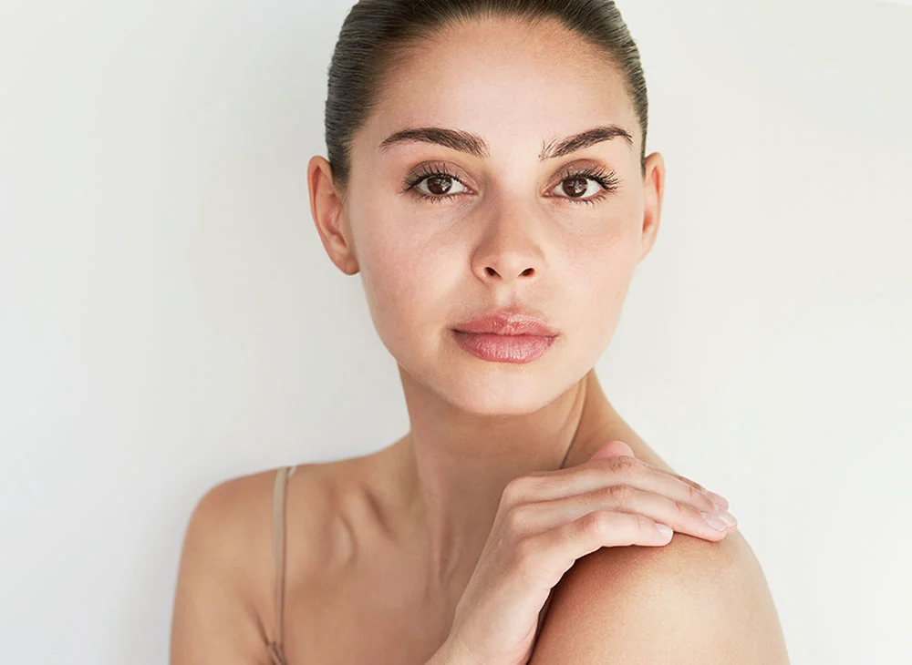 A woman with smooth, light-toned skin faces the camera with a neutral expression. Her hair is pulled back in a bun, and she wears a light beige or tan thin-strap top. The background is a simple light gray or off-white, with soft, even lighting highlighting her features. - Facial Fat Transfer in San Clamente, Aliso Viejo and Rancho Santa Margarita, CA