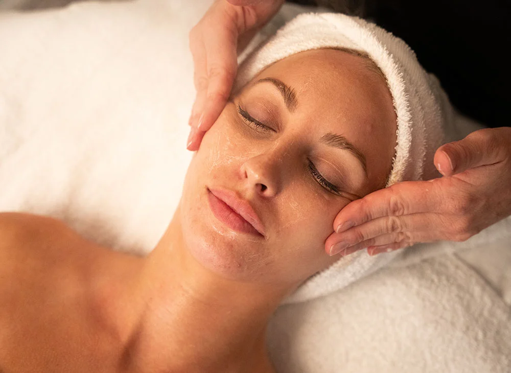A woman lies on a white spa table, eyes closed and expression relaxed, receiving a facial massage. A towel covers her head and neck, while a person’s hands apply pressure to her cheeks and temples. Facial product is visible on her face and the hands performing the massage. Soft lighting focuses on the woman, with a blurred background. - Facials in San Clamente, Aliso Viejo and Rancho Santa Margarita, CA