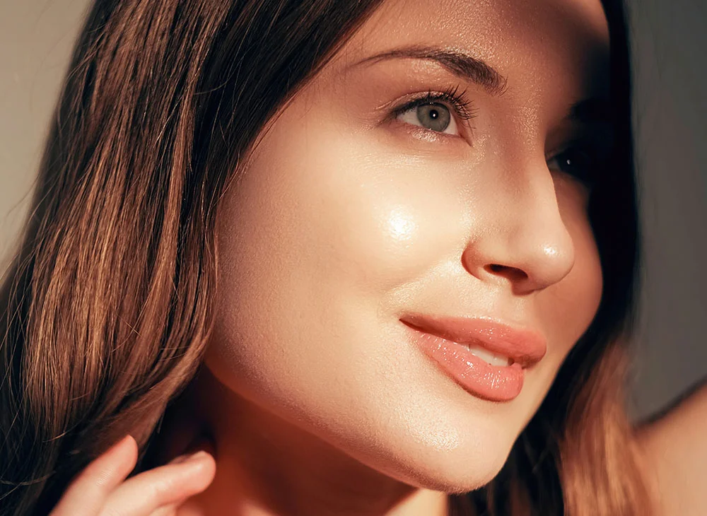 A close-up of a woman with long brown hair and smooth, light-toned skin, gazing slightly to the right. Her lips are a light pinkish-nude, and part of her hand touches her neck and hair. Soft sunlight creates a dramatic interplay of light and shadow, with a blurred, neutral-toned background. - Fractional Co2 Laser in San Clamente, Aliso Viejo and Rancho Santa Margarita, CA