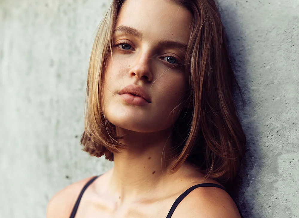 A close-up of a young woman leaning against a textured light gray wall. With shoulder-length, light brown hair and light blue eyes, her expression is calm and composed. Her lightly tanned skin complements the simplicity of her black sleeveless top. Soft, diffused lighting creates a gentle ambiance, subtly blurring the background and drawing attention to her poised presence. - LipLift in San Clamente, Aliso Viejo and Rancho Santa Margarita, CA