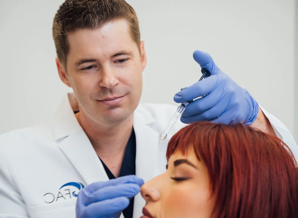 Dr. Bryan Brandon, wearing a white lab coat with a light-blue logo, is applying dermal fillers to a relaxed woman with reddish-brown hair. He wears blue gloves and uses a syringe to treat her forehead. The background is light gray, with even lighting focused on the procedure. - Liquid Rhinoplasty in San Clamente, Aliso Viejo and Rancho Santa Margarita, CA