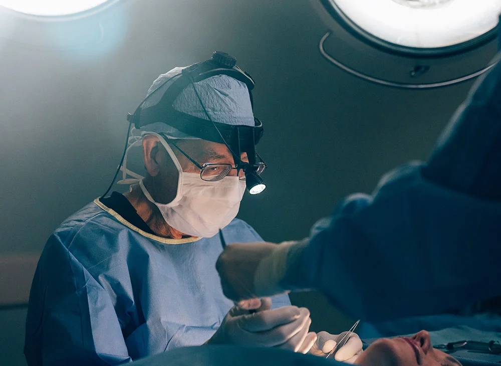 Close-up of Dr. Richard Gangnes performing surgery. He is wearing a surgical gown, mask, and loupe, focused on the patient. A gloved hand from another team member is visible. The operating room is dimly lit, with surgical lights highlighting the scene. - MOHs Reconstruction in San Clamente, Aliso Viejo and Rancho Santa Margarita, CA