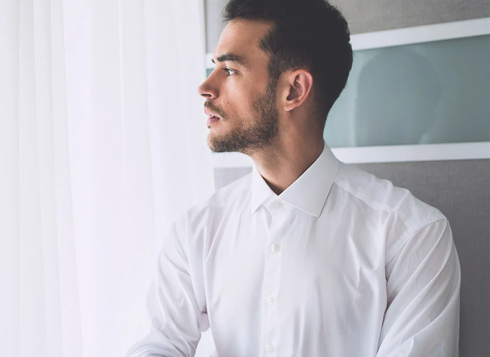 A close-up portrait of a middle-aged man with short dark hair. He wears a white dress shirt and gazes off to the right, appearing thoughtful. The background features a window with sheer curtains and a neutral wall. - PRF Hair Treatment in San Clamente, Aliso Viejo and Rancho Santa Margarita, CA