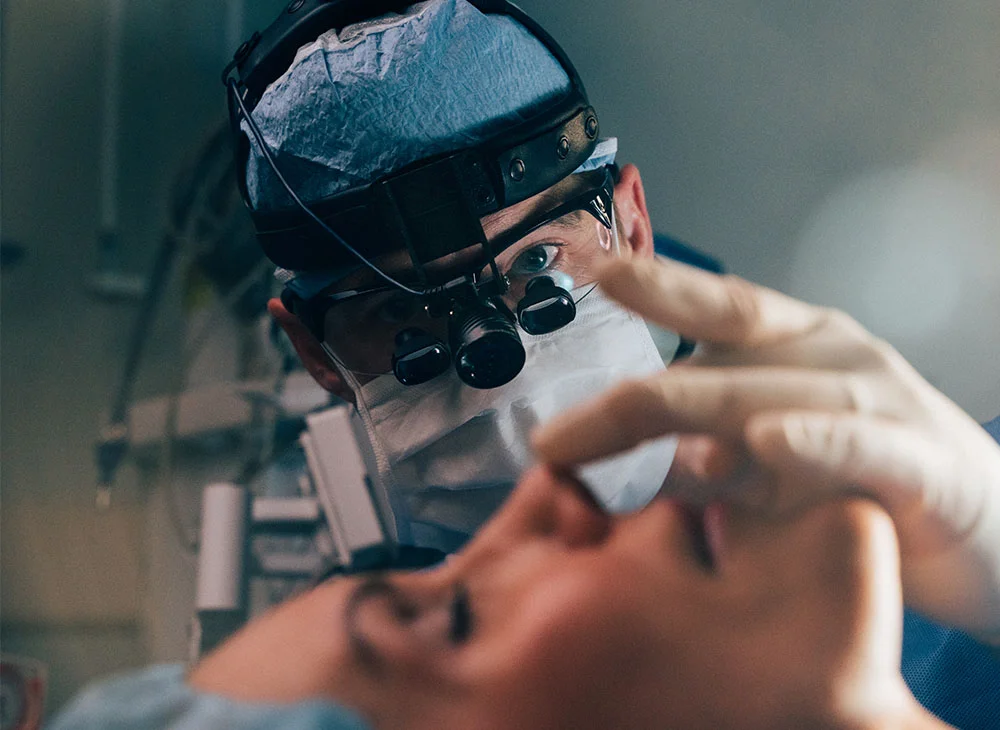 Dr. Brandon, a skilled surgeon, performs a delicate procedure, likely cosmetic or oral surgery. Magnification lenses and precise hand movements are evident as he focuses on the patient's facial area. The clinical setting and advanced equipment underscore the commitment to delivering high-quality care. - Rhinoplasty in San Clamente, Aliso Viejo and Rancho Santa Margarita, CA
